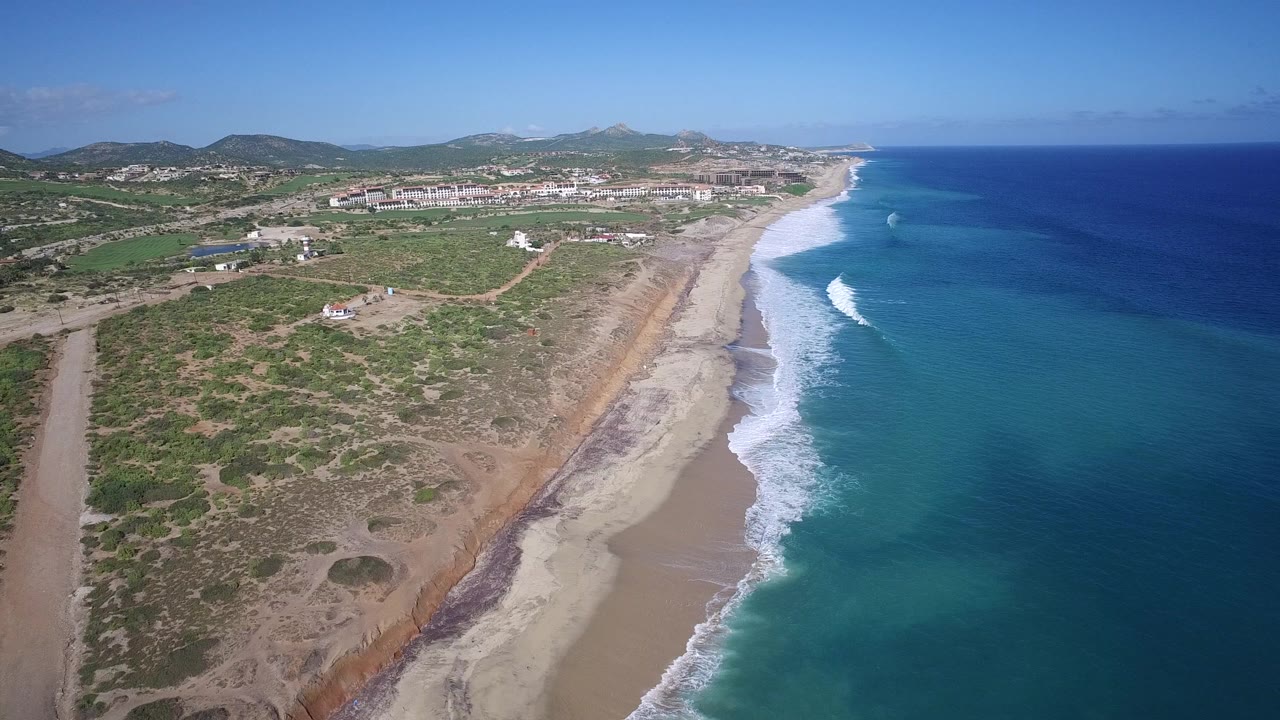 Opening aerial frame over Playita, Baja California Sur, Mexico, with scrubland and dirt roads on the left, a sandy beach through the center, blue ocean on the right, and no visible people.