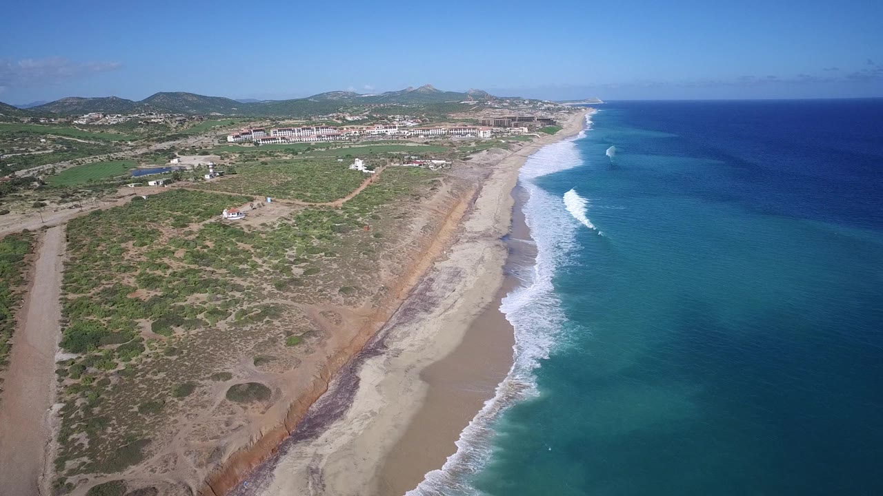 Aerial coastline with blue ocean, surf, sandy beach, scrubland, and low development.