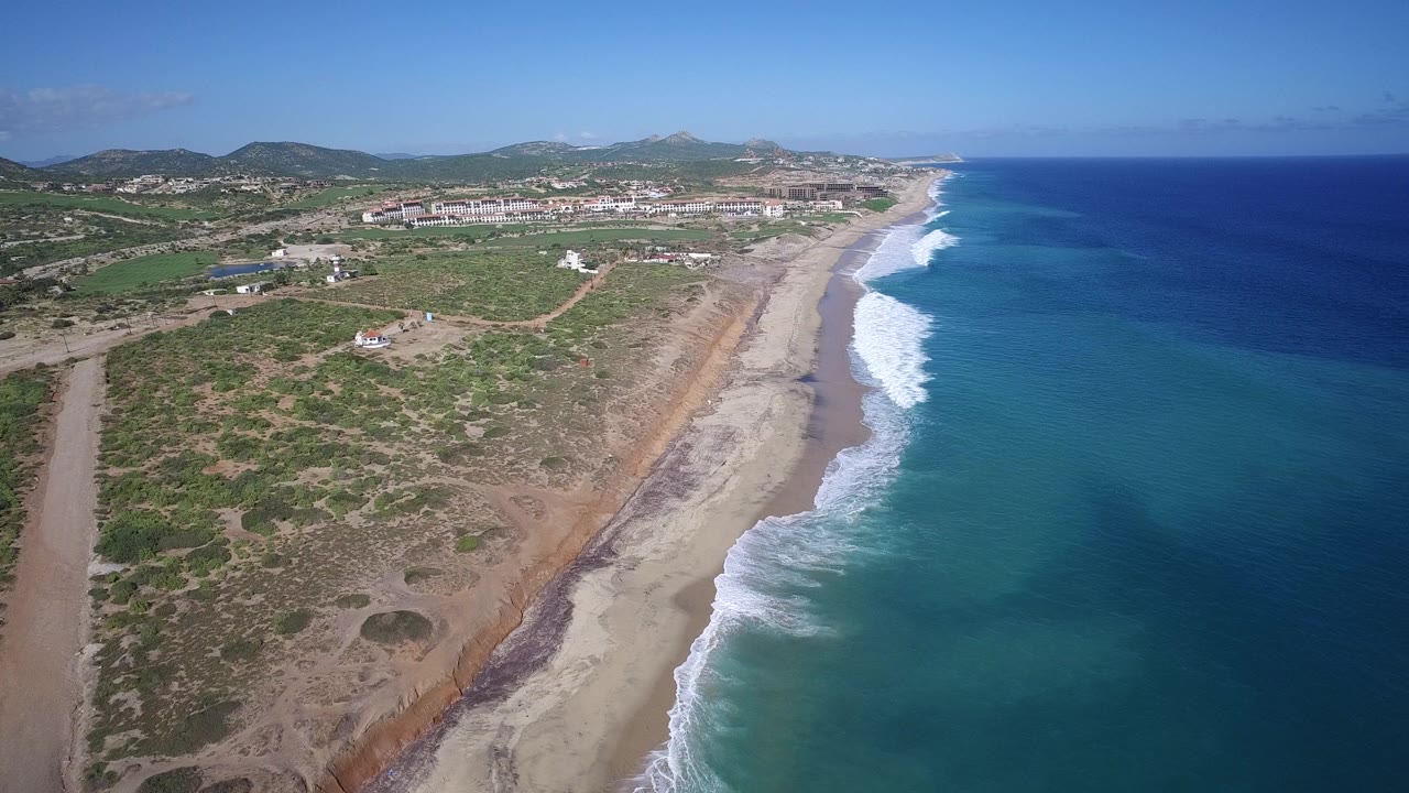 Opening aerial view of Playita, Baja California Sur, Mexico, with blue ocean on the right, white surf along the sand, scrubland and dirt tracks inland, distant hills, and no visible people.