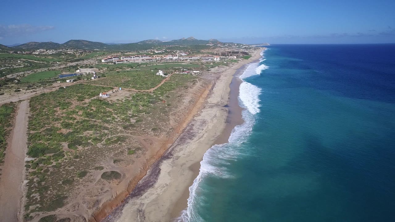 Second aerial frame over Playita, Baja California Sur, Mexico, showing a long sandy beach, turquoise shallows, breaking waves, inland scrub, low buildings, mountains, and no visible people.