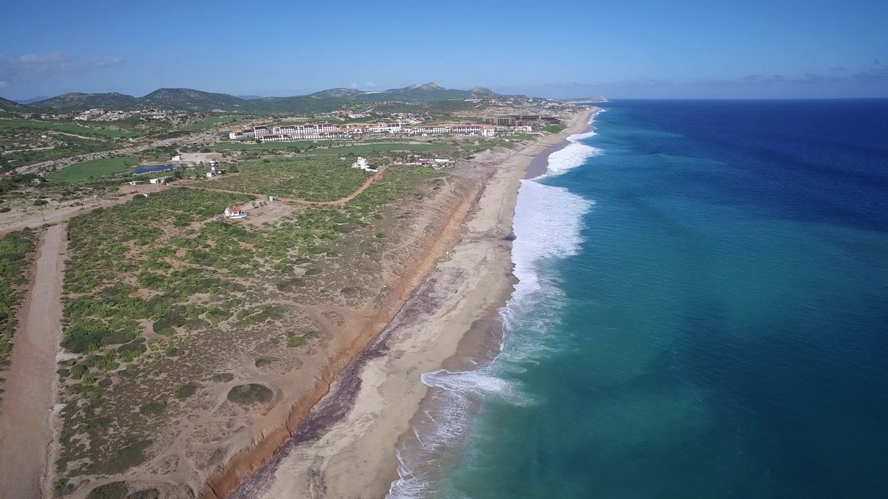 High aerial view over Playita, Baja California Sur, showing sandy beach, white surf, blue-green ocean, scrubland, distant buildings, hills, and no visible people.