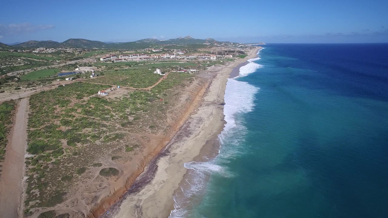 Aerial frame from Playita, Baja California Sur, with the surf line running up the beach, open water on the right, low buildings inland, and no visible people.