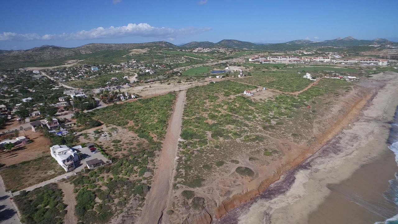 Elevated aerial view in Playita, Baja California Sur showing scrubby land, dirt roads, scattered homes, green fields, distant hills, and no visible people.
