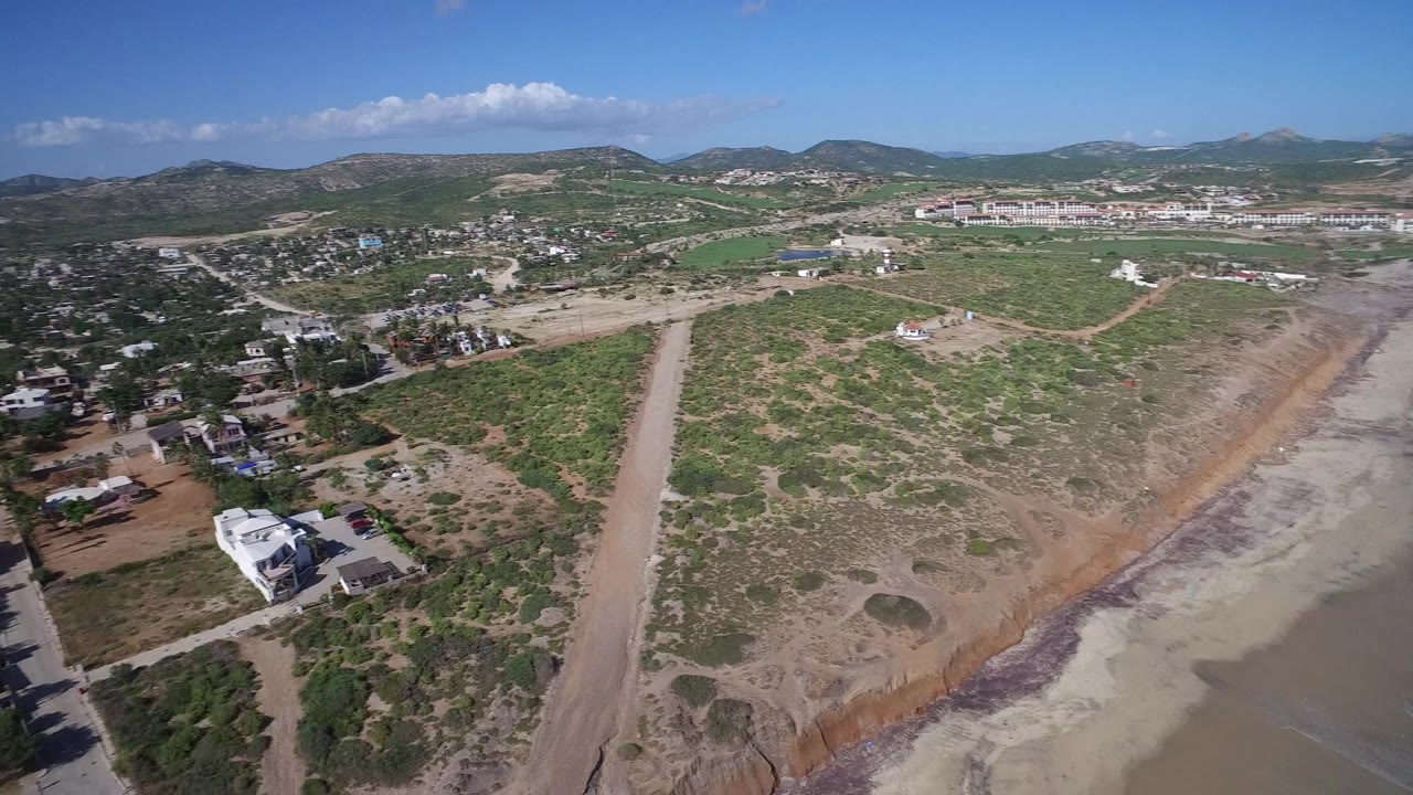Drone-height view in Playita, Baja California Sur with a dirt road, pale water or lagoon channel, scrub vegetation, homes, distant hills, and no visible people.