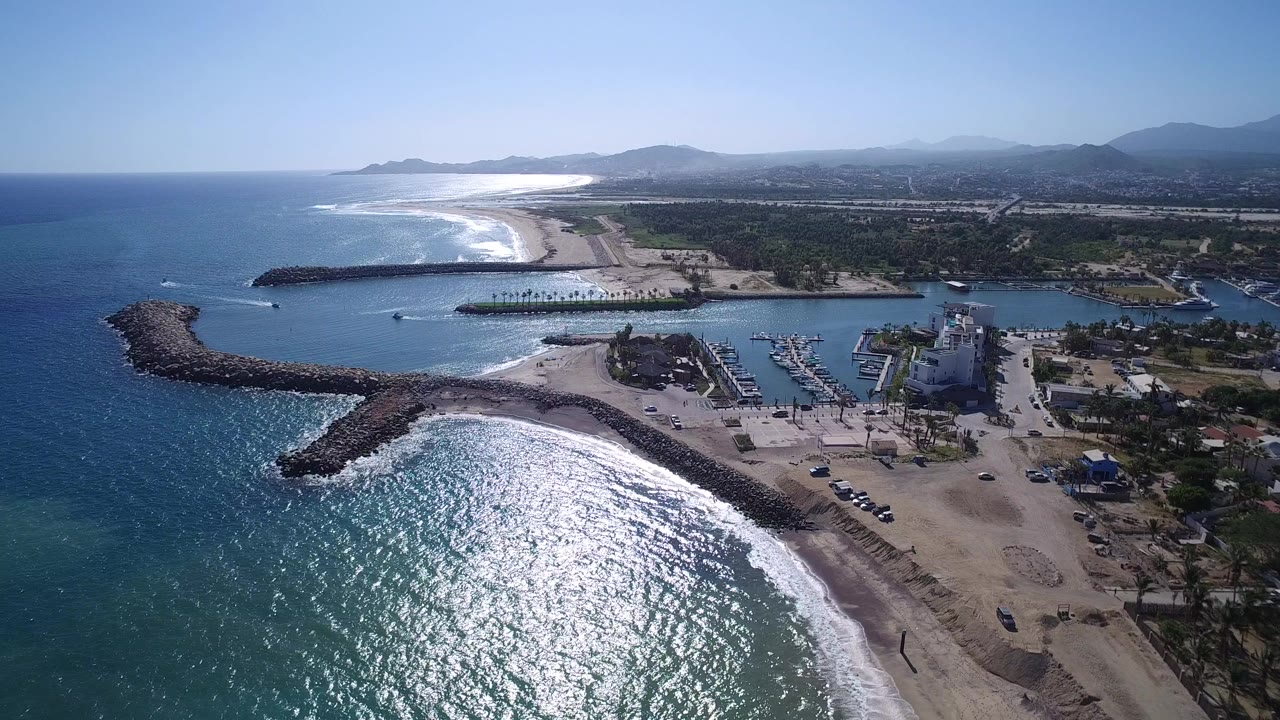 Aerial view of ocean, beach, breakwaters, marina, village, and distant hills.