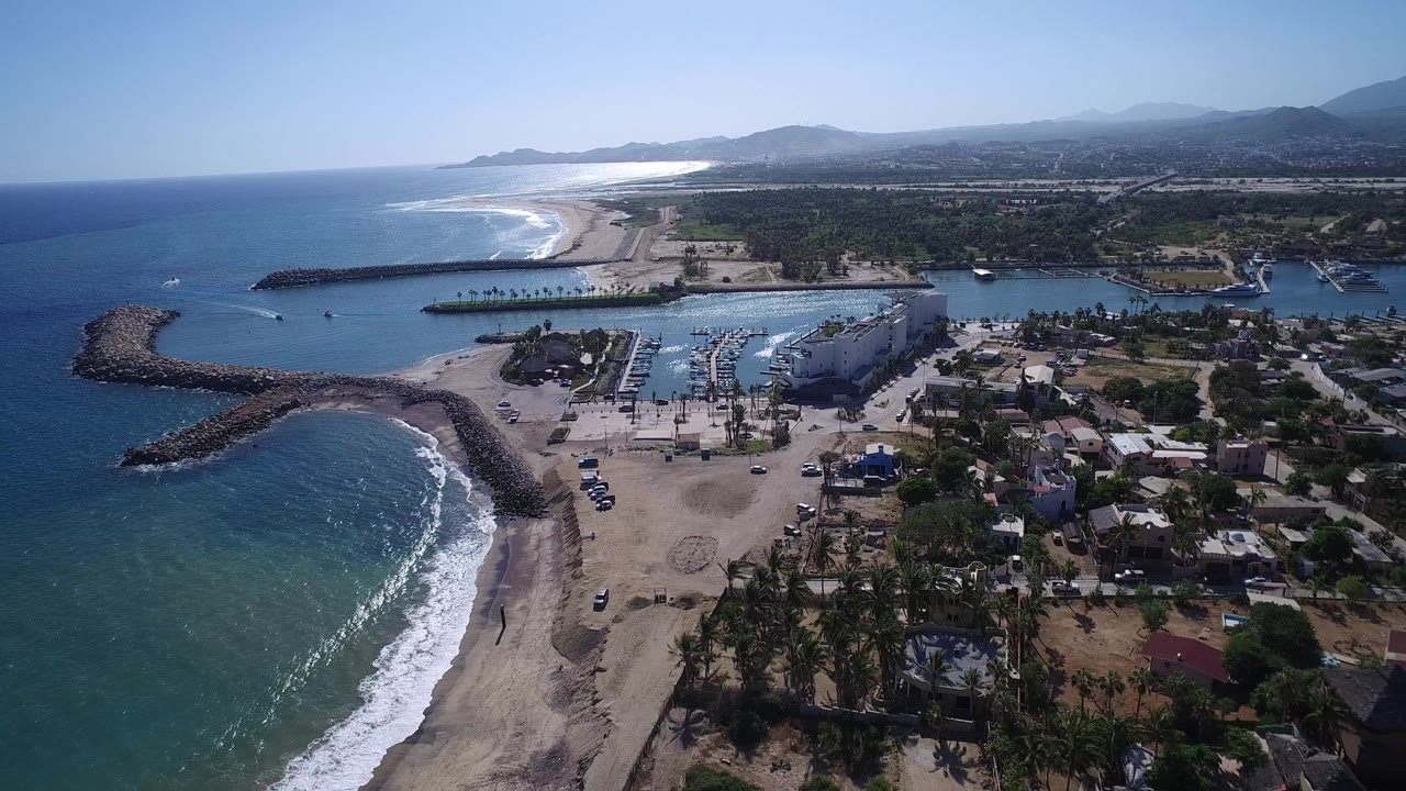 Aerial frame over Playita with the curved beach and white surf on the left, marina slips and boats in the center, low buildings to the right, mountains in the distance, and no visible people.