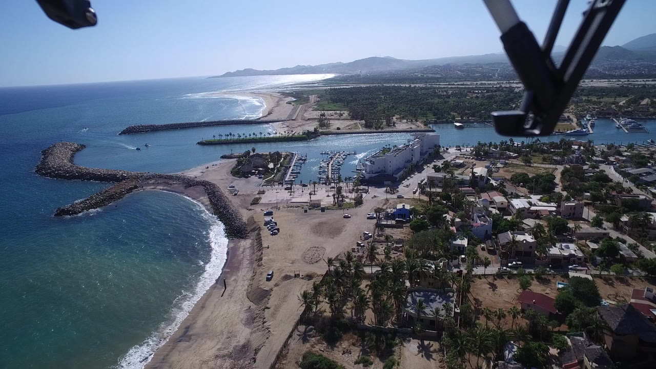 Aerial view of blue ocean, sandy beach, marina, homes, and hills with drone gear at the top edge.