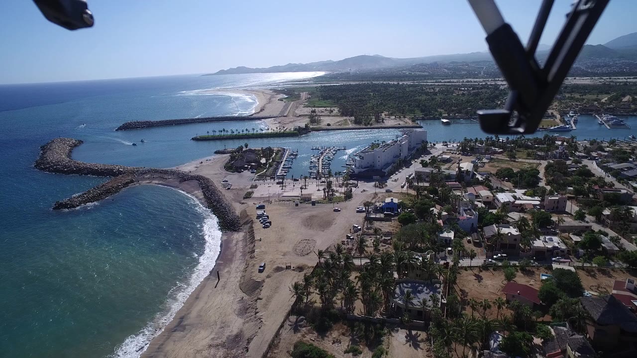 Aerial frame over Playita, Baja California Sur, with the beach and breakwater slightly more centered, marina slips and homes visible inland, and no visible people.