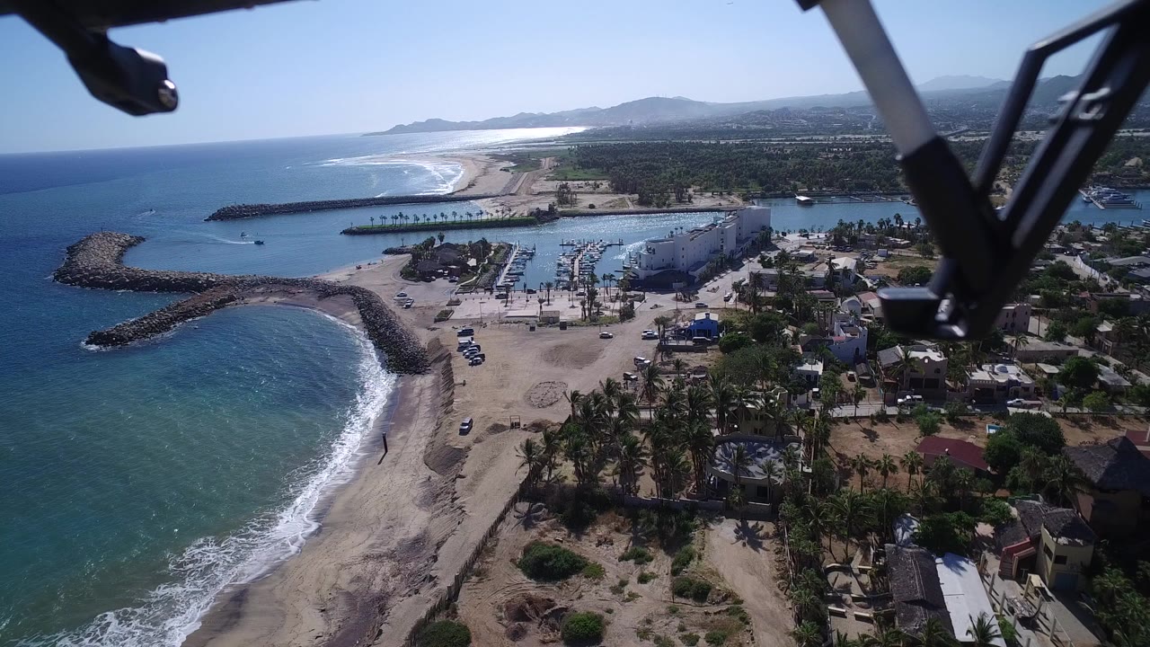 Aerial view over a curved beach, rock breakwaters, turquoise water, and a small harbor.