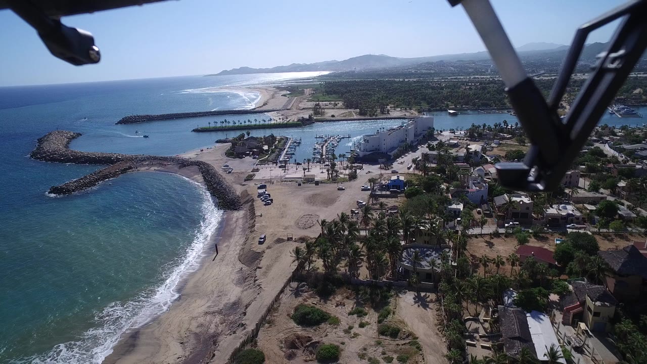 Aerial view at Playita, Baja California Sur, Mexico, with small waves along the beach, a protected harbor to the right, palms and low buildings below, and no visible people.