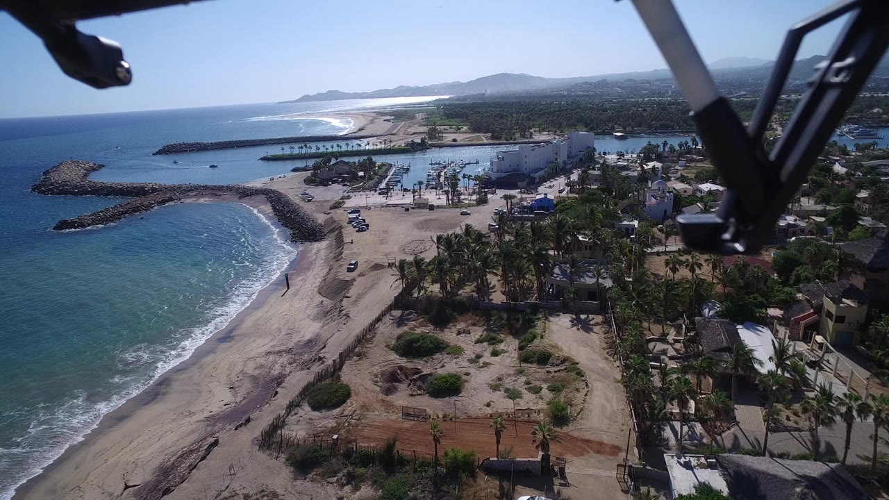 Aerial view of waves breaking along a sandy beach beside palm trees and coastal buildings.