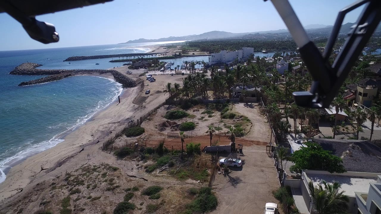Aerial frame of Playita showing the shoreline curving beside blue water, sandy lots and scrub in the foreground, palms and residences to the right, vehicles near a dirt road, small visible people on the beach, and aircraft hardware crossing the upper frame.