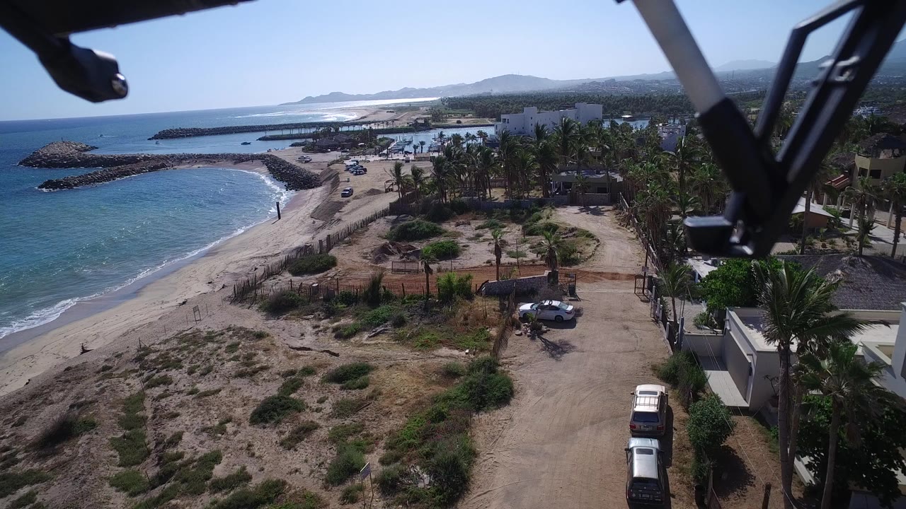 Aerial view in Playita, Baja California Sur with waves curling onto the sand, a light dirt road beside palms and low buildings, parked vehicles, distant hills, and no visible people.