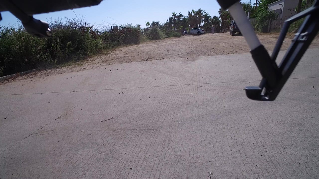 Low ground-level view in Playita, Baja California Sur, with drone landing gear entering the upper frame, bright concrete road below, roadside brush and palms beyond, and no visible people.