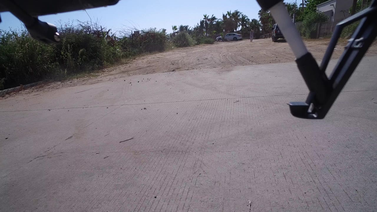 Drone landing gear sits near the top of a low road view in Playita, Baja California Sur, with pale pavement, dry roadside vegetation, distant palms, a parked car, and no visible people.