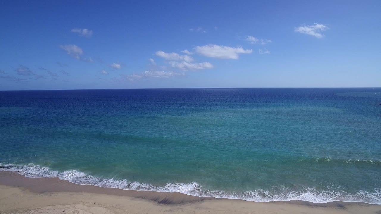 High view of the Playita, Baja California Sur shoreline with turquoise water, a tan sand strip, small breaking waves, blue sky, scattered clouds, and no visible people.
