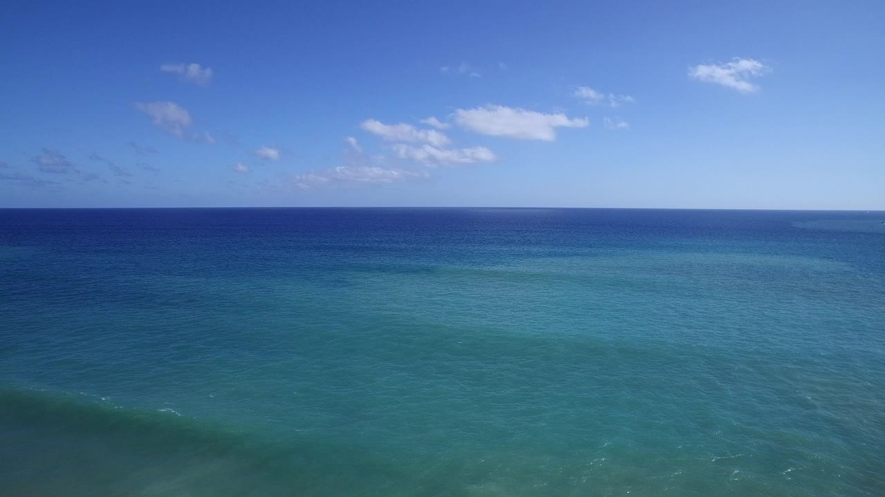 Opening aerial view over blue-green sea in Playita, Baja California Sur, Mexico, with a dark blue horizon band, scattered clouds, and no visible people.