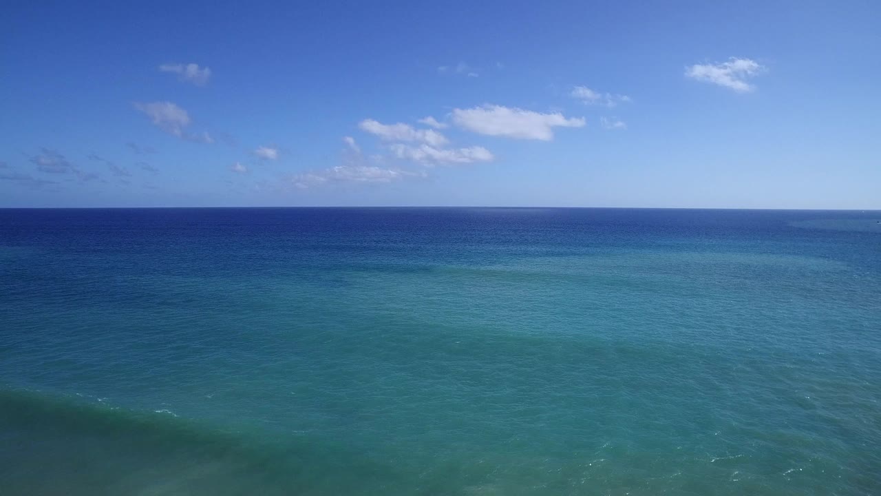 Aerial frame over Playita blue-green water with a broad turquoise foreground, deeper blue sea near the horizon, small clouds overhead, and no visible people.