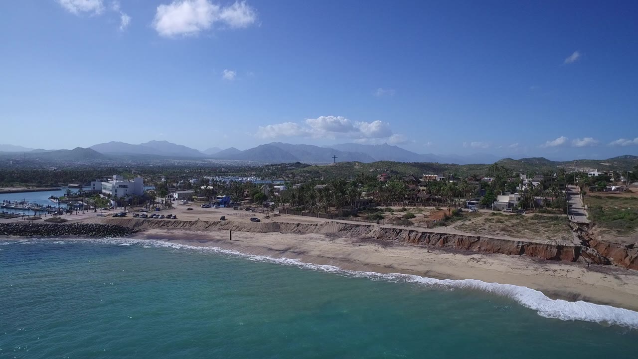 Aerial view of turquoise surf, sandy beach, coastal buildings, trees, and distant mountains.