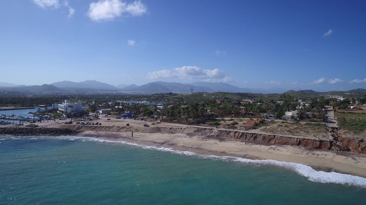 Wide aerial frame of Playita shoreline, showing blue-green ocean at lower left, a line of breaking waves, the sandy beach, clustered buildings, trees, hills, and no visible people.