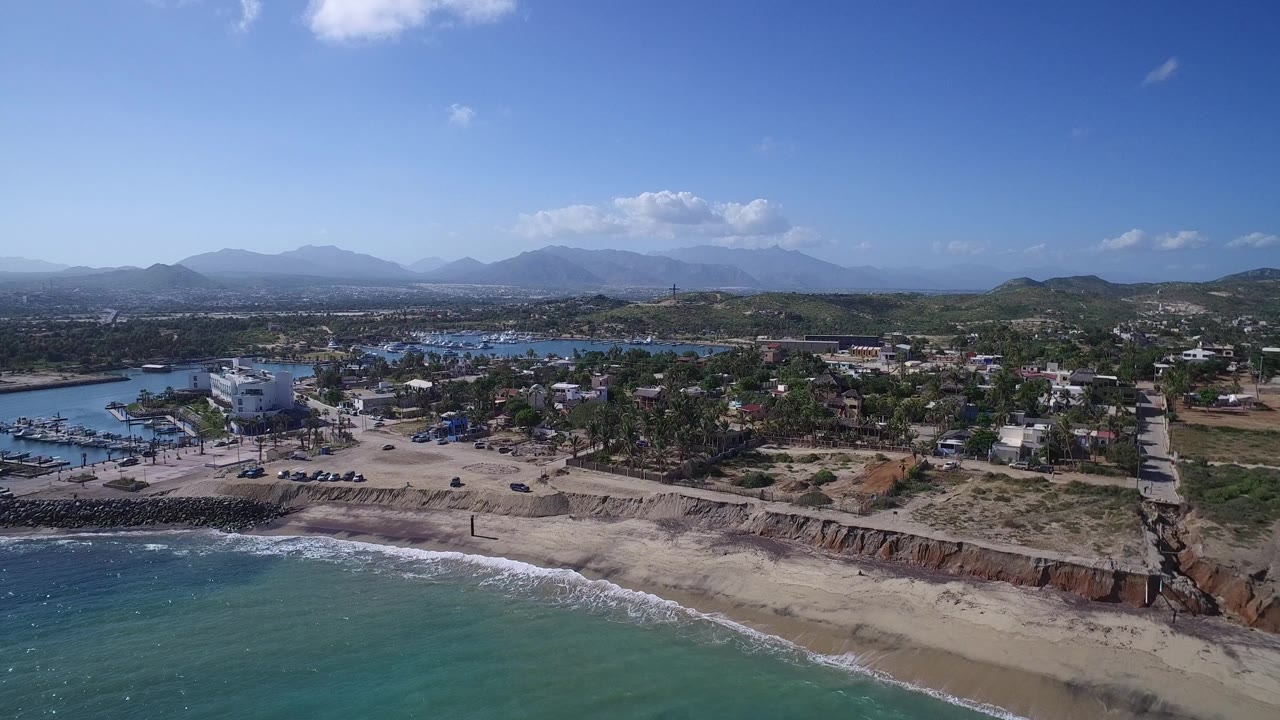 Drone-height view of Playita with surf crossing the lower frame, a pale beach and bluff, marina water to the left, low buildings inland, mountains on the horizon, and no visible people.