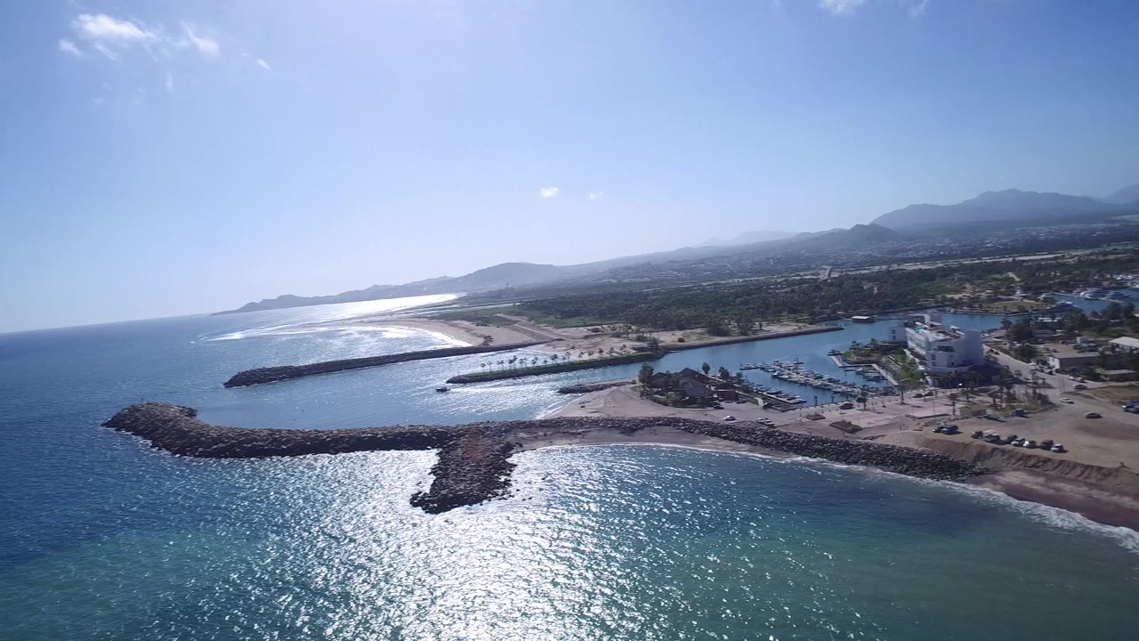 Wide aerial frame in Playita, Baja California Sur, Mexico with the ocean in the foreground, rock breakwaters around harbor water, a sandy shore, low buildings inland, distant hills, and no visible people.
