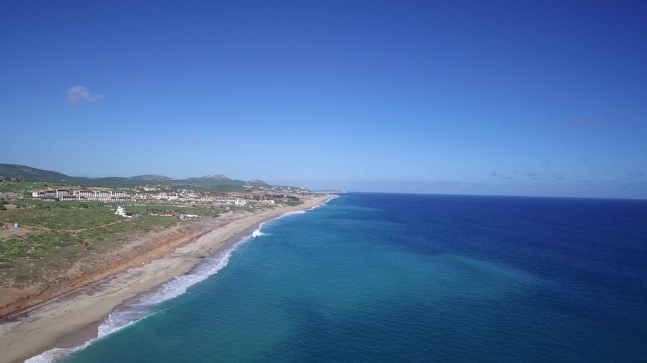 Aerial view over the Playita, Baja California Sur coastline with deep blue ocean, turquoise shallows, a sandy beach, distant buildings, and no visible people.