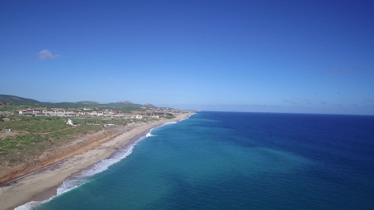 Wide aerial view of surf meeting a long sandy beach beside coastal scrubland.