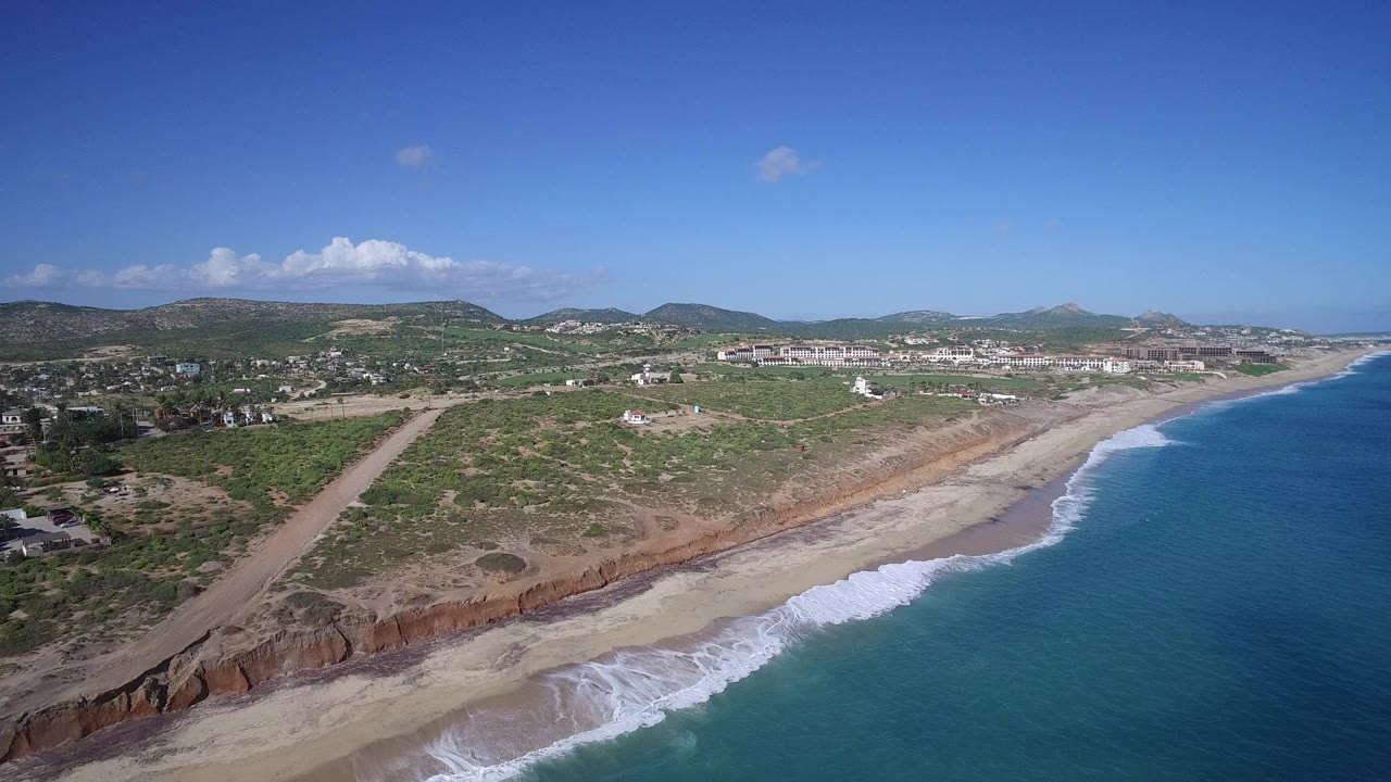 Opening aerial view over Playita, Baja California Sur, with blue ocean on the right, white surf along a sandy beach, reddish bluffs, inland scrubland, low mountains, and no visible people.