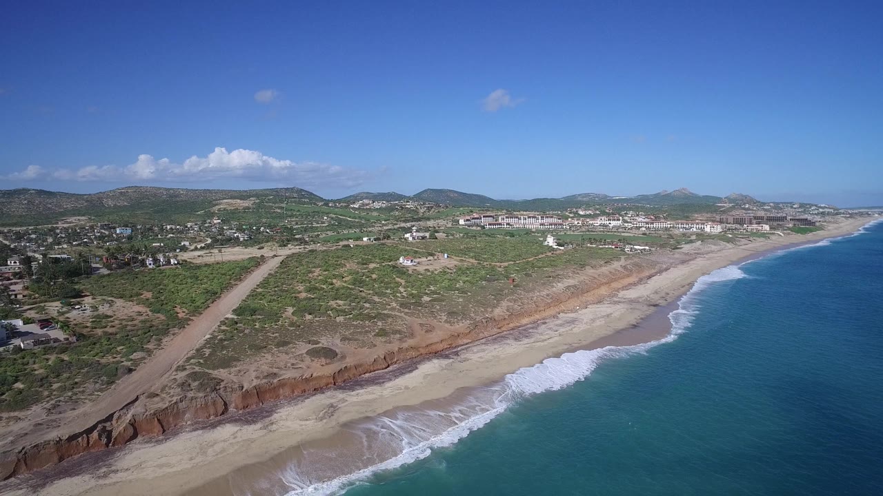 Drone view of beach, ocean, inland scrub, buildings, and mountains under clear sky.