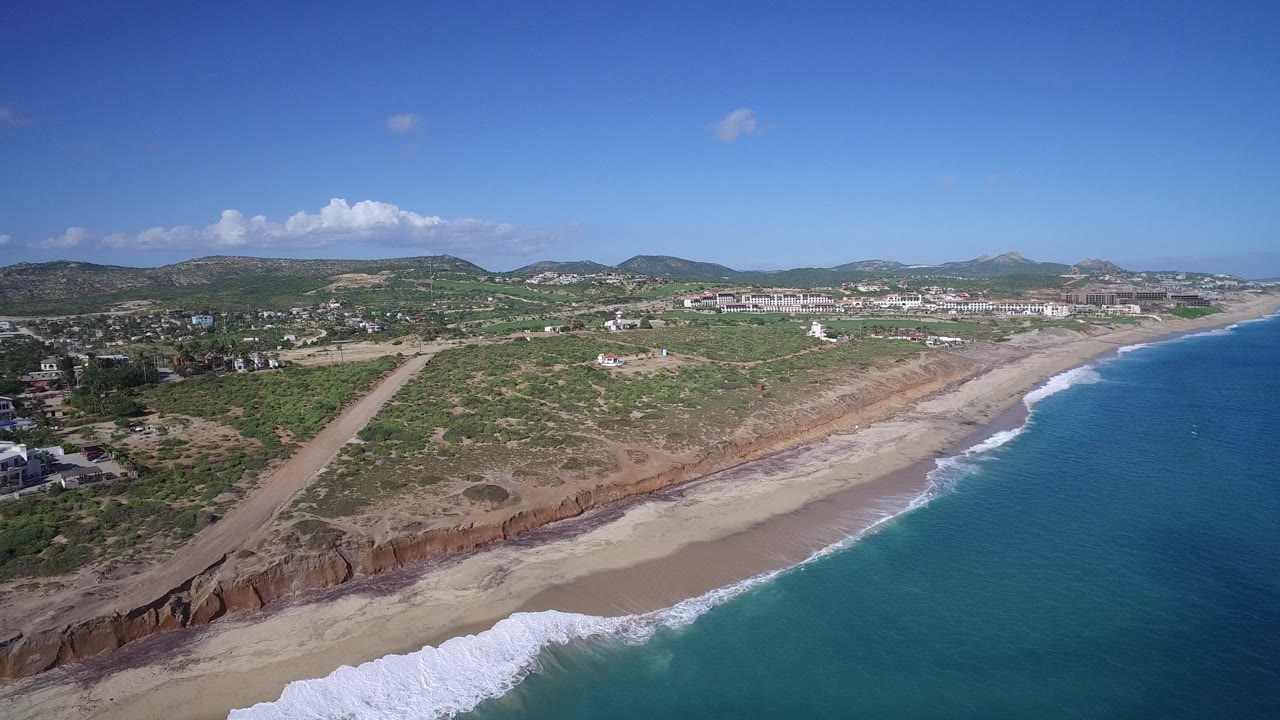 Aerial view over Playita, Baja California Sur, with white surf reaching a sandy beach below reddish bluffs, dirt roads and low hills inland, and no visible people.