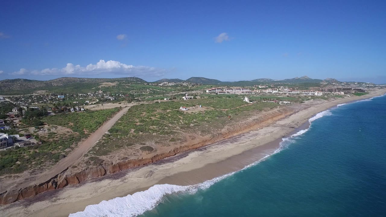 Drone frame over Playita showing surf bands along the beach, a diagonal dirt road through scrubland, scattered buildings at left, distant mountains, and no visible people.