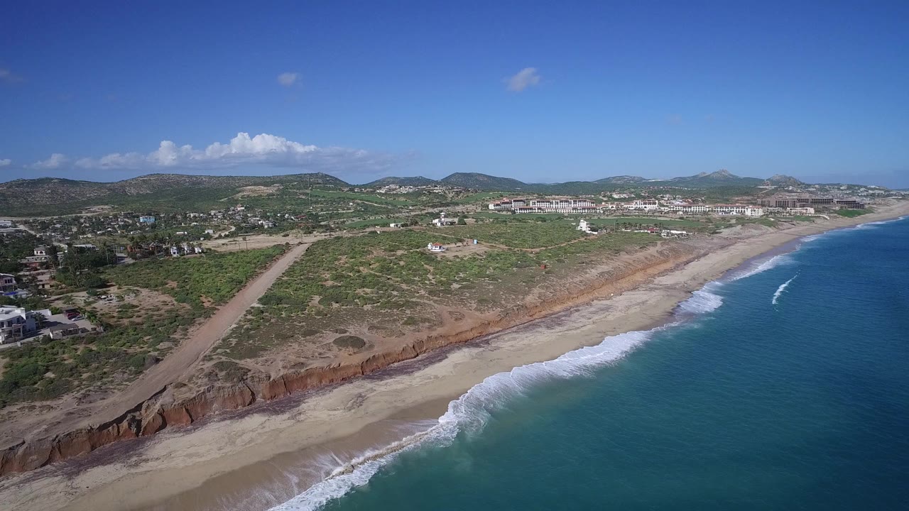 Aerial view in Playita, Baja California Sur, with the shoreline running diagonally, a fresh white wave line near the beach, dry scrubland inland, houses at the left edge, hills on the horizon, and no visible people.