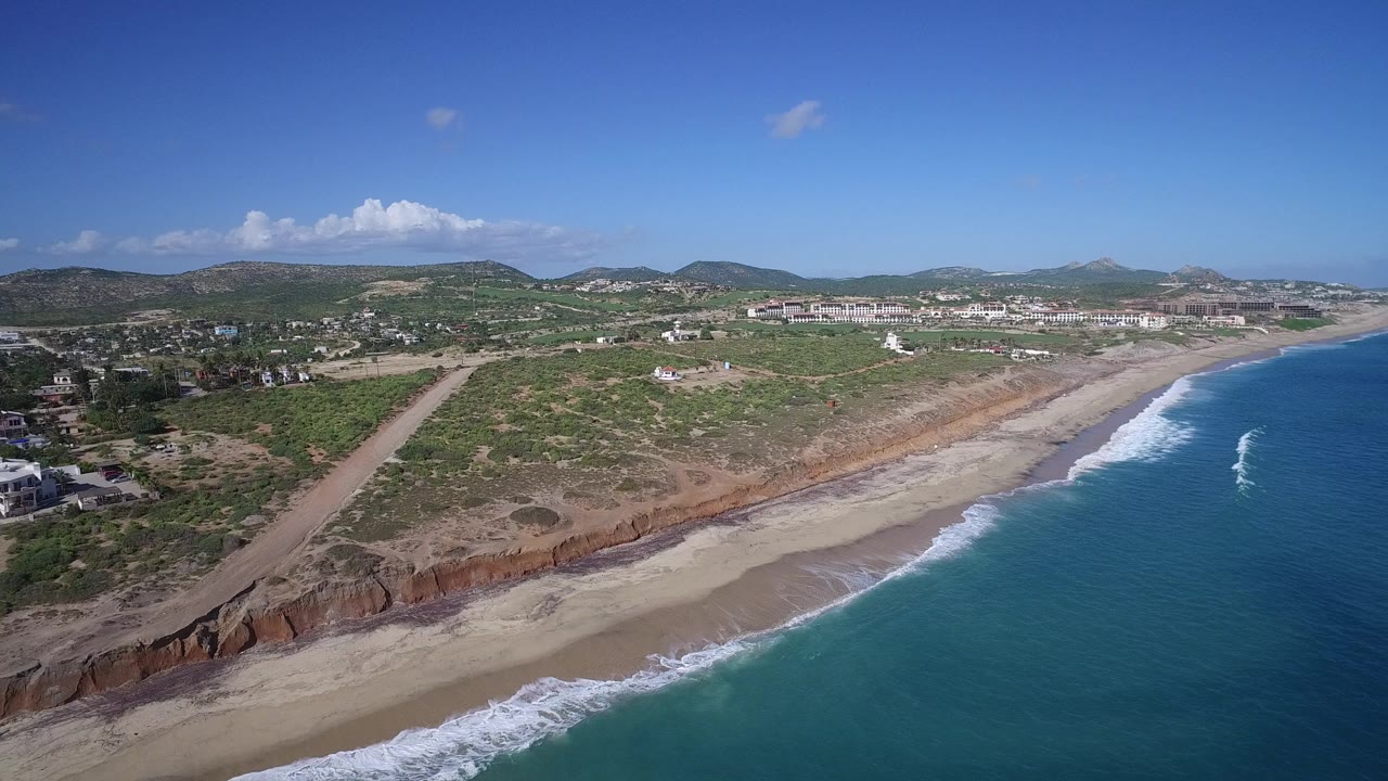 Aerial view over Playita, Baja California Sur showing blue ocean, a sandy beach, reddish coastal bluffs, scrubland, scattered buildings, distant hills, and no visible people.
