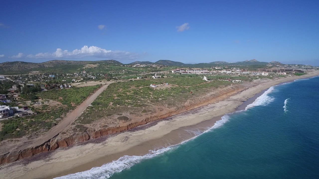 Wide aerial frame of Playita, Baja California Sur with the ocean filling the right side, surf reaching the sand, scrubland beyond the bluff, distant buildings, and no visible people.