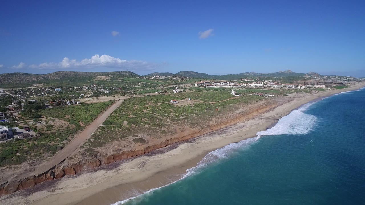 Aerial view of surf, beach, shrub-land, roads, houses, and hills under blue sky.