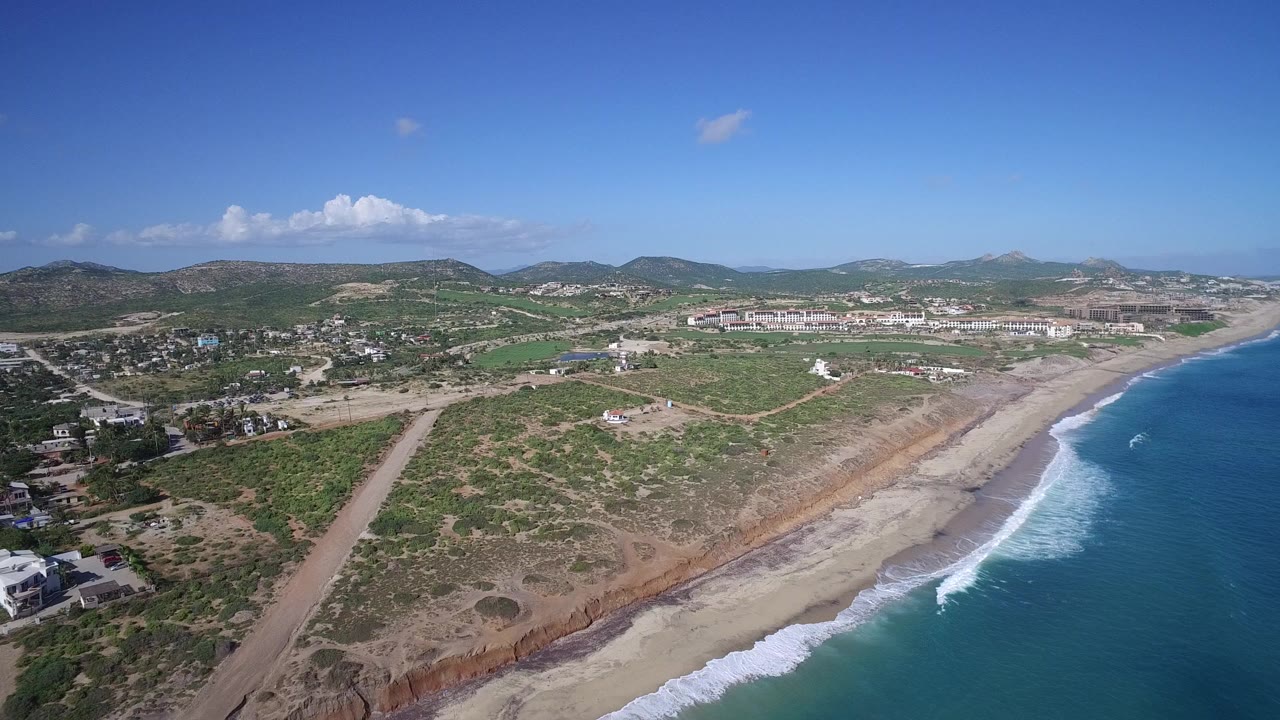 High aerial view of Playita, Baja California Sur with blue ocean on the right, surf along the beach, scrubland and roads inland, distant hills, and no visible people.