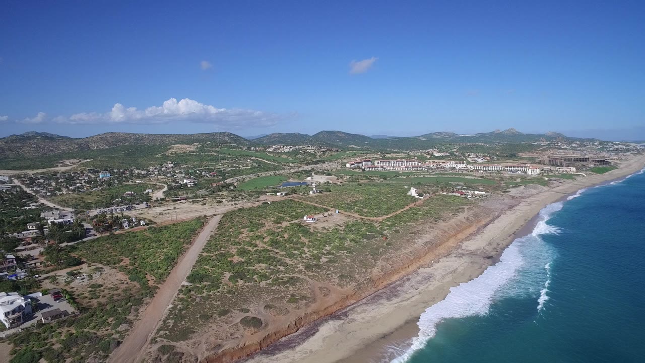 Opening aerial frame over Playita, Baja California Sur, with blue surf along the right edge, pale beach below, scrubland and dirt roads inland, scattered homes, distant hills, and no visible people.