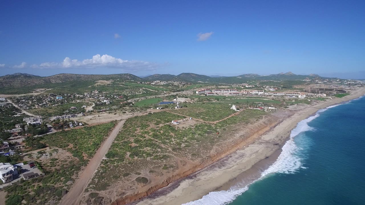 Aerial frame over Playita, Baja California Sur, showing blue ocean at right, white surf on the beach, scrubland, roads, scattered buildings, distant hills, and no visible people.