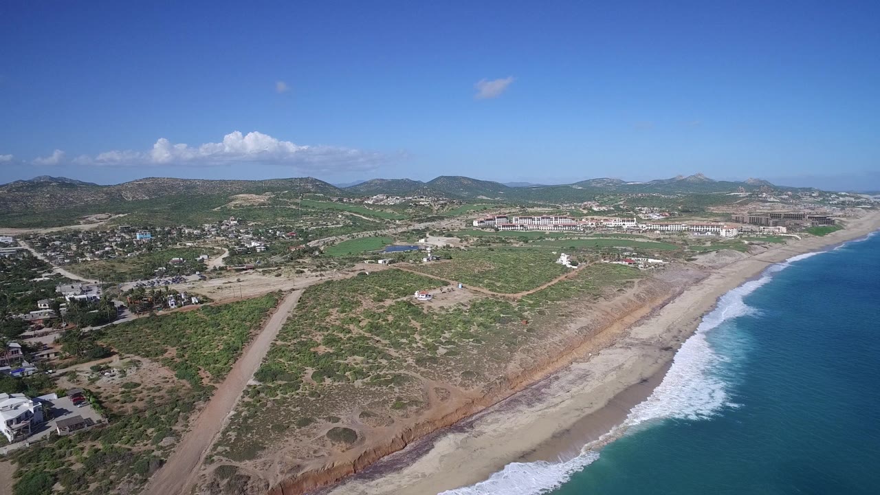 High coastal aerial with waves, beach, open scrubland, roads, and inland buildings.