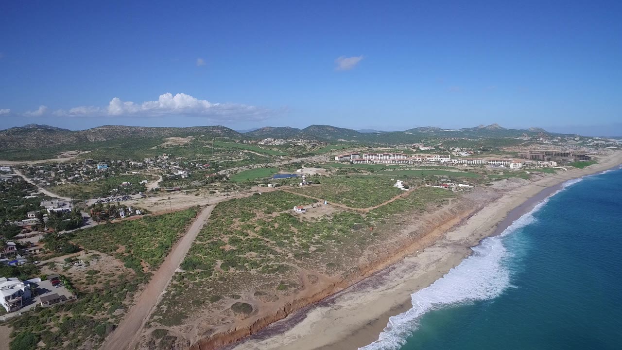 High aerial frame over Playita, Baja California Sur with blue ocean at right, white surf on the sandy beach, scrubland and dirt roads inland, low mountains on the horizon, and no visible people.