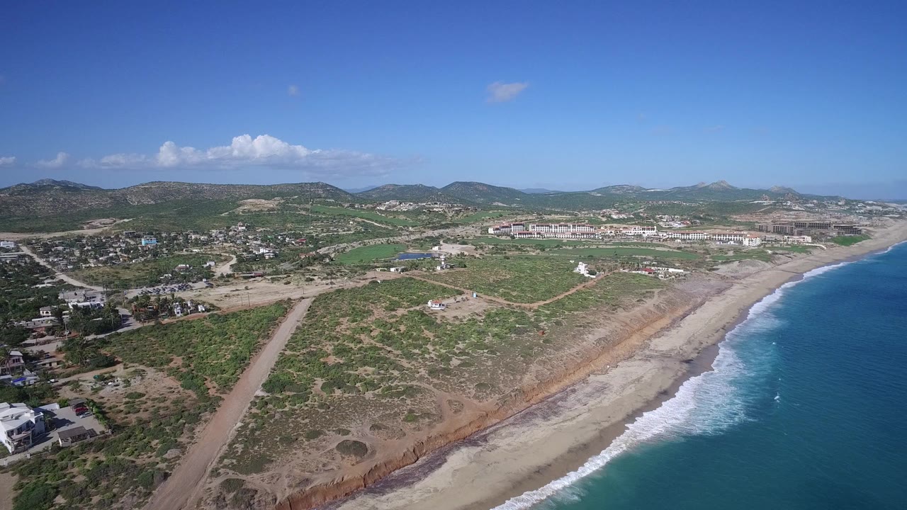 Aerial frame over Playita with the ocean on the right, surf meeting a pale beach, dry lots and roads through the center, houses and green patches inland, distant mountains, and no visible people.