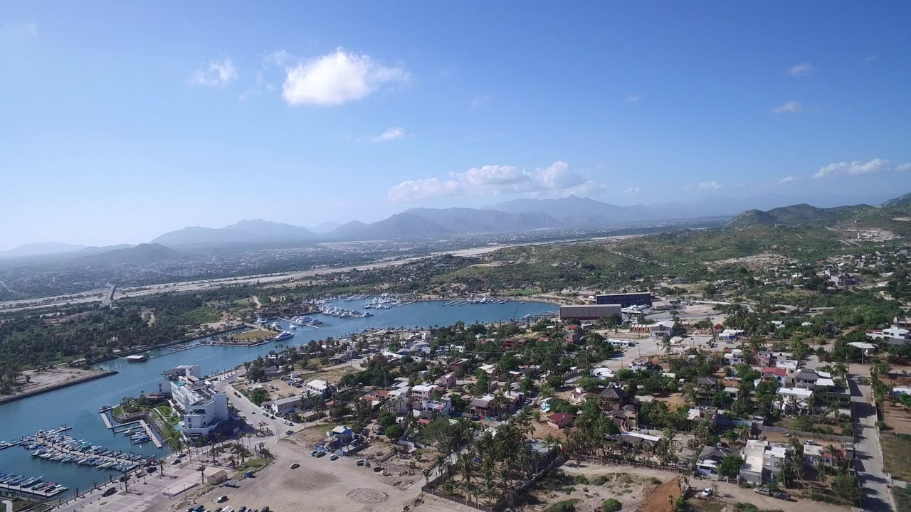 Opening frame of Playita, Baja California Sur from high above, with marina water on the left, low buildings and trees below, dry hills beyond, and hazy mountains under a blue sky.