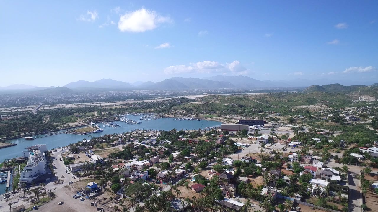 At 0:01, the Playita aerial holds on a broad view of residential blocks, a road corridor, blue harbor water with moored boats, green-brown hills, and hazy mountains beyond.
