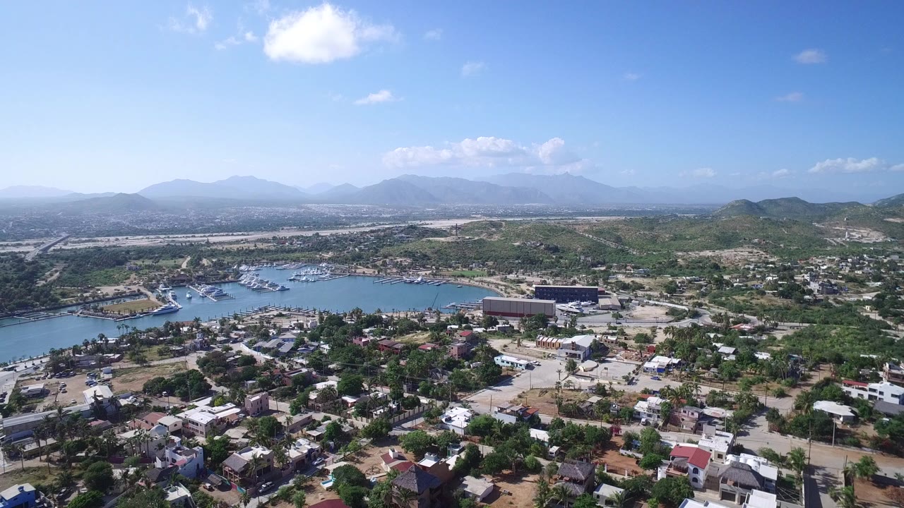 Aerial view of a coastal town, blue water, boats, hills, and distant mountains.