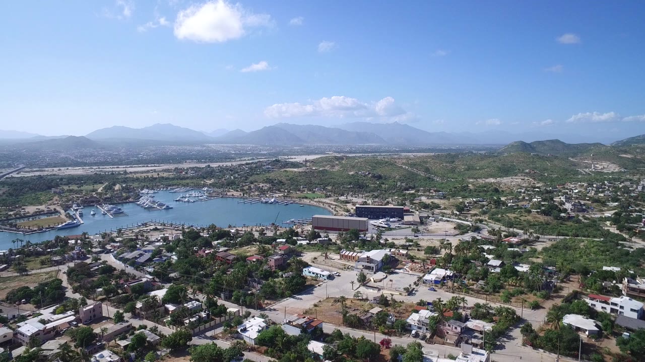 Aerial view over a waterfront town, blue water, boats, hills, and distant mountains.
