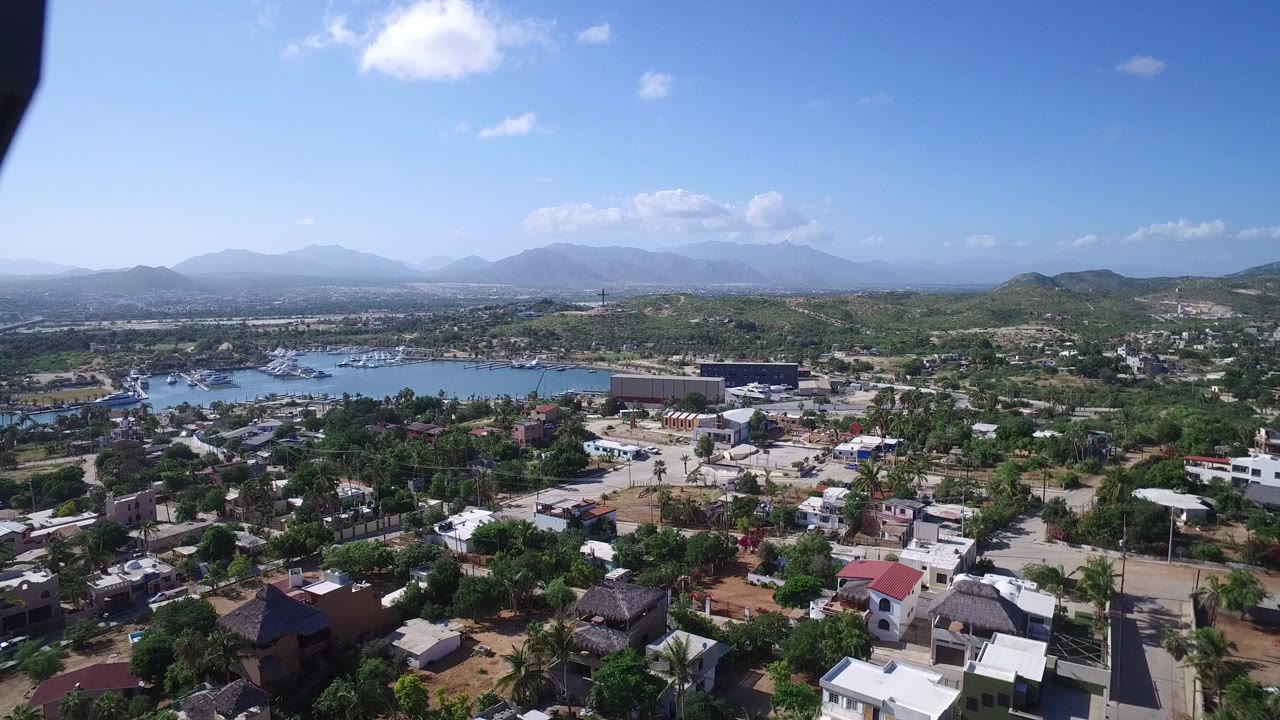 Aerial frame over Playita, Baja California Sur with clustered houses and greenery in the foreground, water across the midground, mountains on the horizon, and no visible people.