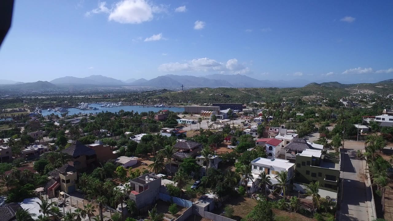 Early aerial view in Playita with clustered homes and trees in the foreground, a blue inlet across the middle distance, and mountains under scattered clouds, with no visible people.