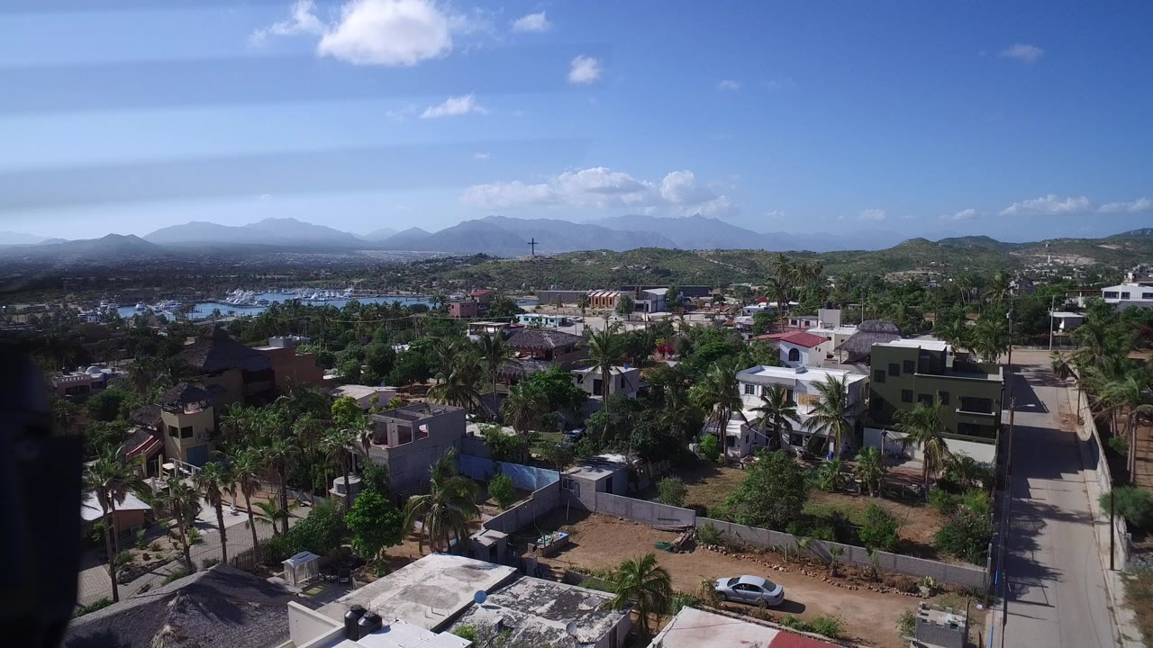 High aerial view over Playita, Baja California Sur, showing low homes, palms, roads, blue water beyond the town, distant hills, scattered clouds, and no visible people.