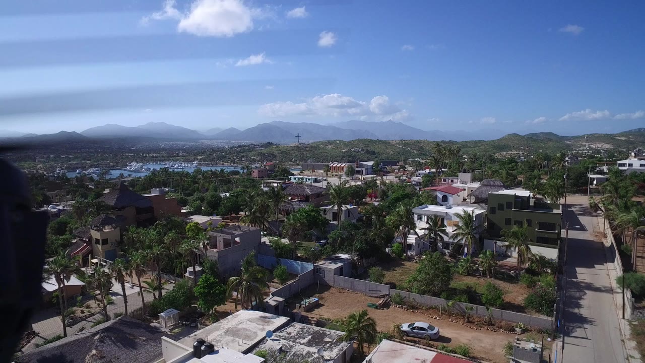 Aerial view over Playita with houses, palms, roads, distant water, hazy hills, blue sky, and a dark aircraft or drone component along the left edge, with no visible people.